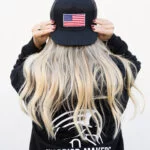 Back view of a woman wearing the black Warrior Makers classic trucker hat with an American flag patch and a WARRIOR MAKERS t-shirt
