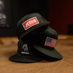 Two black Warrior Makers classic trucker hats stacked on a wood table, one with a red JESUS patch and one with the white Spartan helmet logo