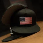 Black Warrior Makers classic trucker hat with an American flag patch on the front panel, shown on a wood table