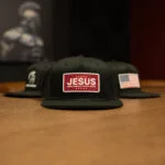 Three black Warrior Makers classic trucker hats lined up on a wood table, one with the Spartan helmet logo, one with a JESUS patch and one with an American flag patch