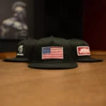 Black Warrior Makers classic trucker hat with an American flag patch, between a Spartan helmet hat and a JESUS patch hat on a wood table