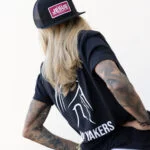Woman wearing the black Warrior Makers classic trucker hat with a pink JESUS patch, seen from behind in a Warrior Makers t-shirt