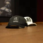 Two Warrior Makers foam trucker hats on a wood table, one olive and one cream, each with a Spartan helmet patch