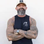 Bearded man with arms crossed wearing the black Warrior Makers sleeveless t-shirt and a cream foam trucker hat