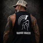 Back view of a tattooed man in a tan hat wearing a black Warrior Makers muscle tank with Spartan helmet logo in front of a dark Spartan helmet backdrop
