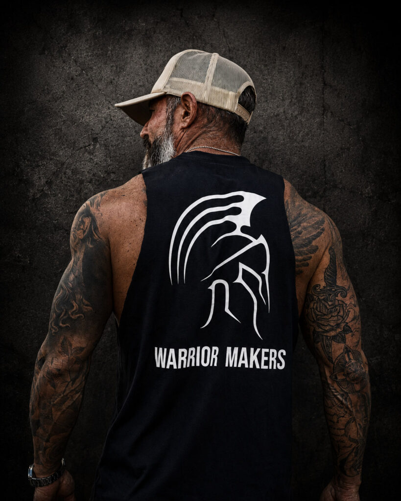 Back view of a tattooed man in a tan hat wearing a black Warrior Makers muscle tank with Spartan helmet logo in front of a dark Spartan helmet backdrop