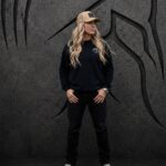 Woman with long blonde hair wearing a black Warrior Makers crewneck sweatshirt and tan hat in front of a dark Spartan helmet backdrop