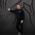 Man in a black Warrior Makers crewneck sweatshirt pointing to the side in front of a dark Spartan helmet backdrop