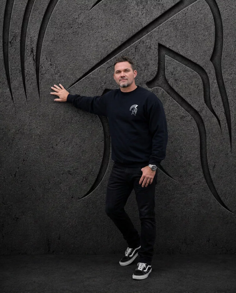 Man in a black Warrior Makers crewneck sweatshirt pointing to the side in front of a dark Spartan helmet backdrop