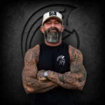 Bearded tattooed man with arms crossed wearing a black Warrior Makers tank top and white trucker hat in front of a dark Spartan helmet backdrop