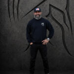 Man wearing a black Warrior Makers crewneck sweatshirt, black pants and hat in front of a dark Spartan helmet backdrop