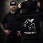 Back view of a man wearing a medium Warrior Makers black hoodie with white Spartan helmet logo in front of a dark Spartan helmet backdrop