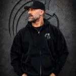 Close-up of a man wearing an XXL Warrior Makers black zip-up hoodie with chest logo and black hat