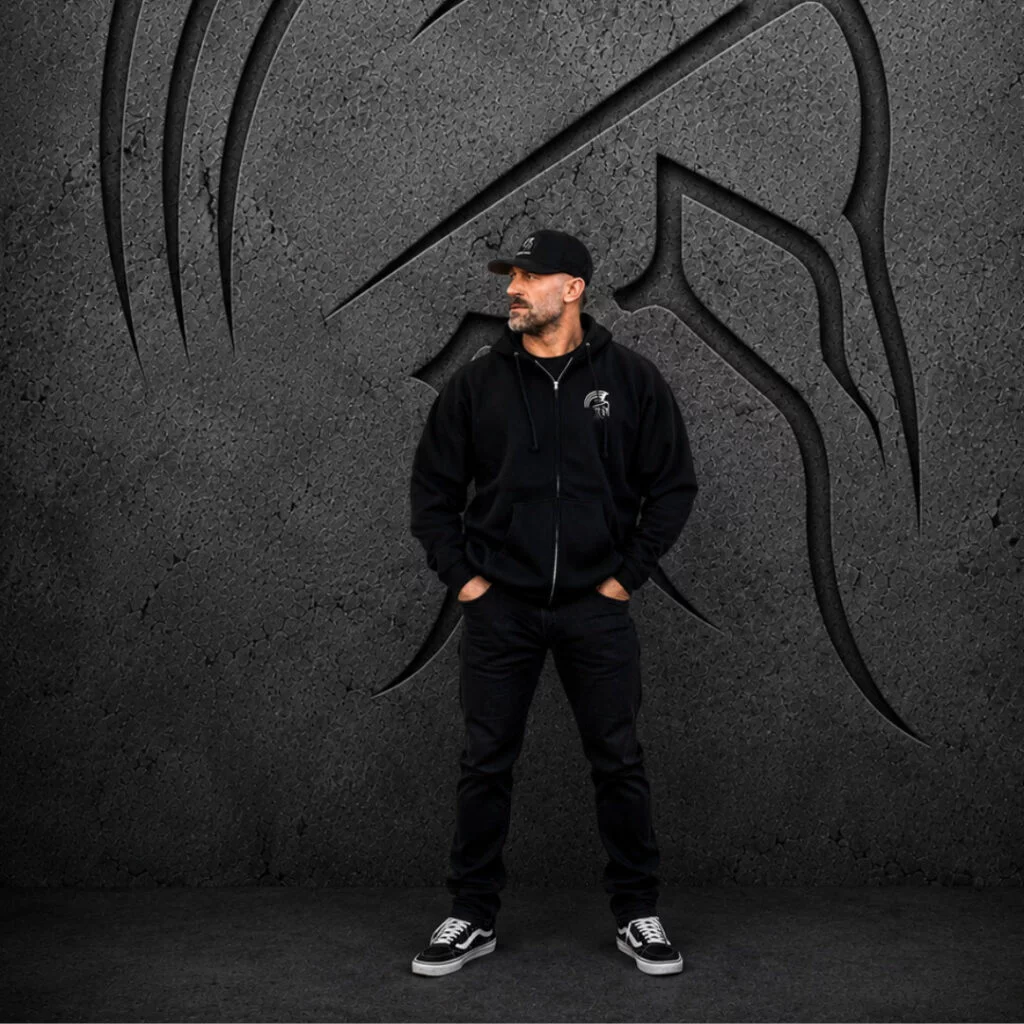 Man wearing an XXL Warrior Makers black zip-up hoodie, black pants and hat in front of a dark Spartan helmet backdrop