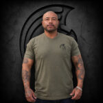 Man wearing an XL Warrior Makers military green t-shirt with small chest logo, standing in front of a dark Spartan helmet backdrop