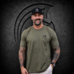 Bearded man wearing an XXL Warrior Makers military green t-shirt with small chest logo, standing in front of a dark Spartan helmet backdrop and black hat