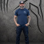 Man wearing a large Warrior Makers navy blue t-shirt with small chest logo, standing in front of a dark Spartan helmet backdrop and black hat