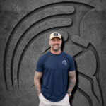Man wearing an XXL Warrior Makers navy blue t-shirt and tan trucker hat in front of a dark Spartan helmet backdrop