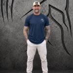 Man wearing an XXL Warrior Makers navy blue t-shirt, khaki pants and tan hat in front of a dark Spartan helmet backdrop