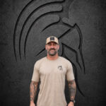 Man wearing a sand tan Warrior Makers t-shirt and tan trucker hat in front of a dark Spartan helmet backdrop