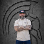 Bearded man with arms crossed wearing a large Warrior Makers tan t-shirt and black hat in front of a dark Spartan helmet backdrop