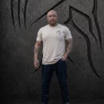 Man wearing an XL Warrior Makers tan t-shirt with small chest logo in front of a dark Spartan helmet backdrop