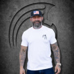 Man wearing a large Warrior Makers white t-shirt and black hat with American flag patch in front of a dark Spartan helmet backdrop
