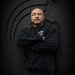 Man with arms crossed wearing an XL Warrior Makers black crewneck sweatshirt in front of a dark Spartan helmet backdrop